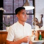 Casual Asian man in a white shirt holding a coffee mug, standing in a kitchen setting.